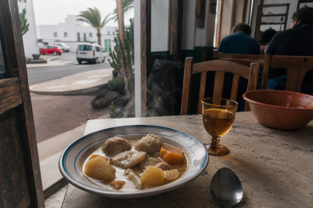 A bowl of salt fish stew with potatoes, sweet potato and gofio dumplings beside a glass of amber wine