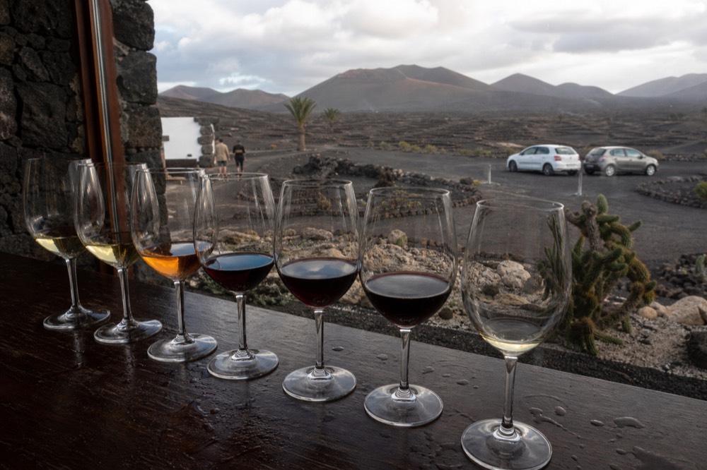 A row of wine glasses on a dark wooden tasting counter with black volcanic ash visible through the window behind