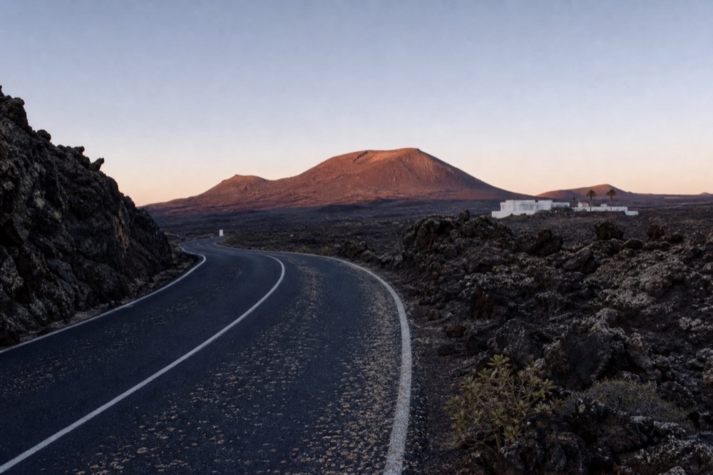 An empty asphalt road curving between black ash slopes with a volcano on the horizon